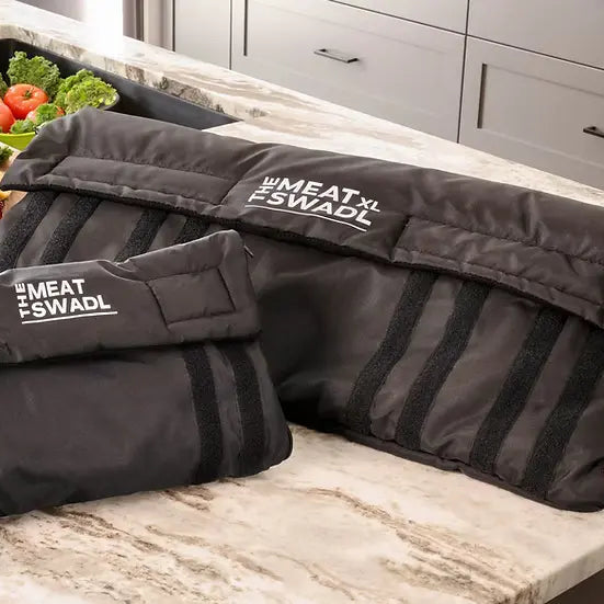 Two black Meat Swadl bags on a kitchen counter with a roasted turkey and vegetables in the background.