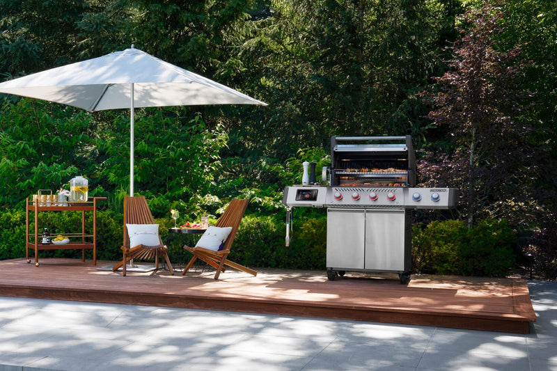 Outdoor patio setup by a swimming pool with a large white umbrella, two wooden lounge chairs, a small side table, and a stainless steel gas grill on a raised wooden deck, surrounded by dense green trees.