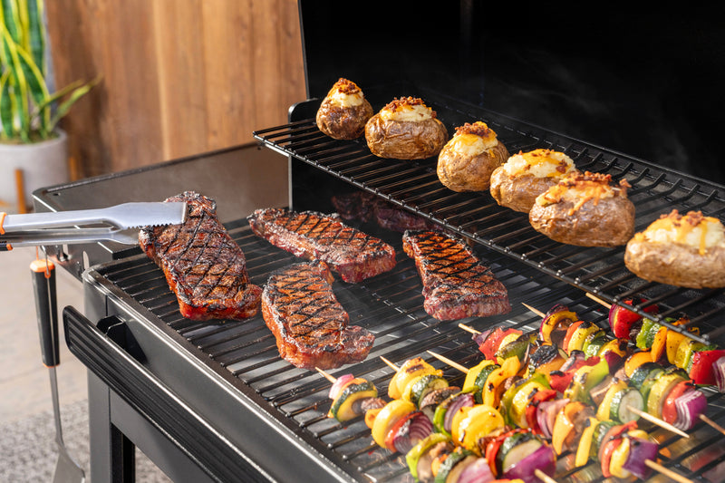 A black outdoor grill loaded with cooked steaks, baked potatoes, and colorful vegetable skewers, with metal tongs resting on the left side.
