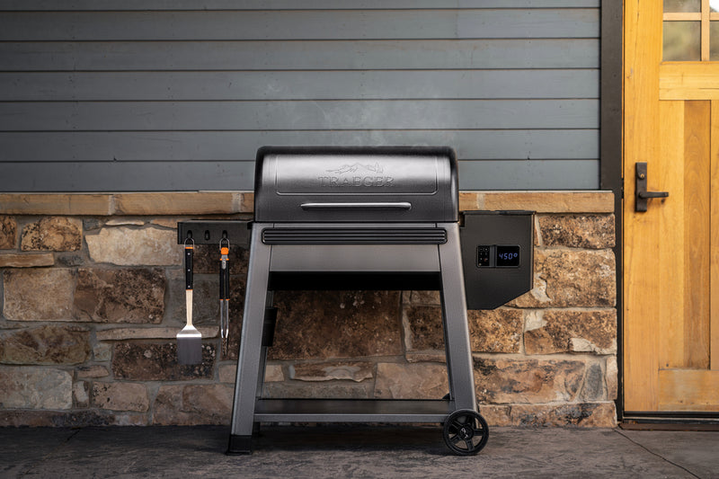 A charcoal grill with a lid, shown outdoors against a gray wood-paneled wall and stone base, with a wooden door on the right. A side-mounted digital controller and hanging utensils are visible.