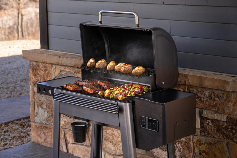 A black outdoor pellet grill with the lid open, cooking meat, potatoes, and vegetables on a patio in front of a gray house wall and stone foundation.