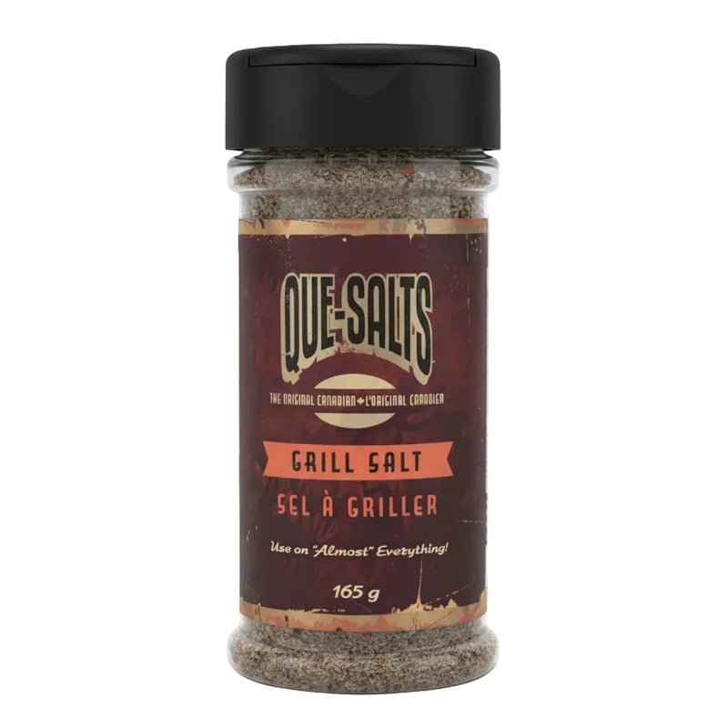 A bottle of Que Salts Grill Salt on a white background. The product is a clear plastic shaker bottle filled with a dark, speckled seasoning blend, topped with a black flip-top lid.